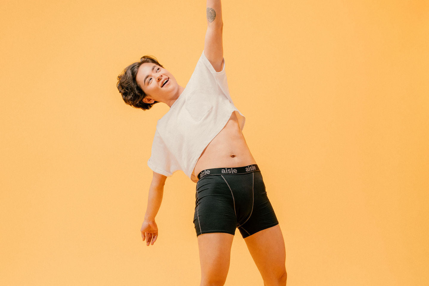 A white model with short brown hair is standing, reaching up to the sky with a smile on their face while wearing a pair of Aisle Boxer Brief period underwear and a white shirt.