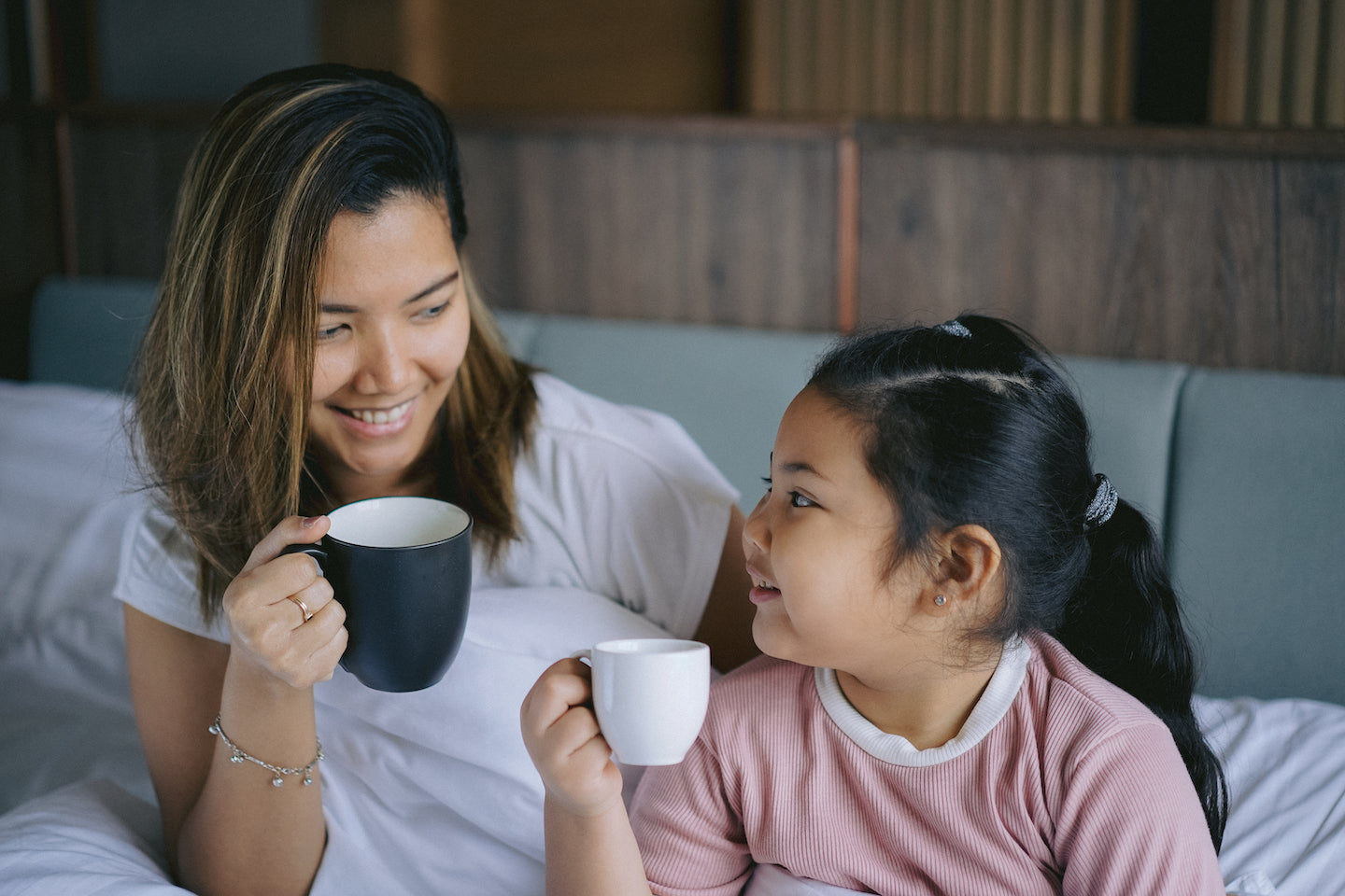 A mother in a white T-shirt holding a black mug sitting with her daughter on a bed. They're looking at each other and smiling, as if mid-conversation.