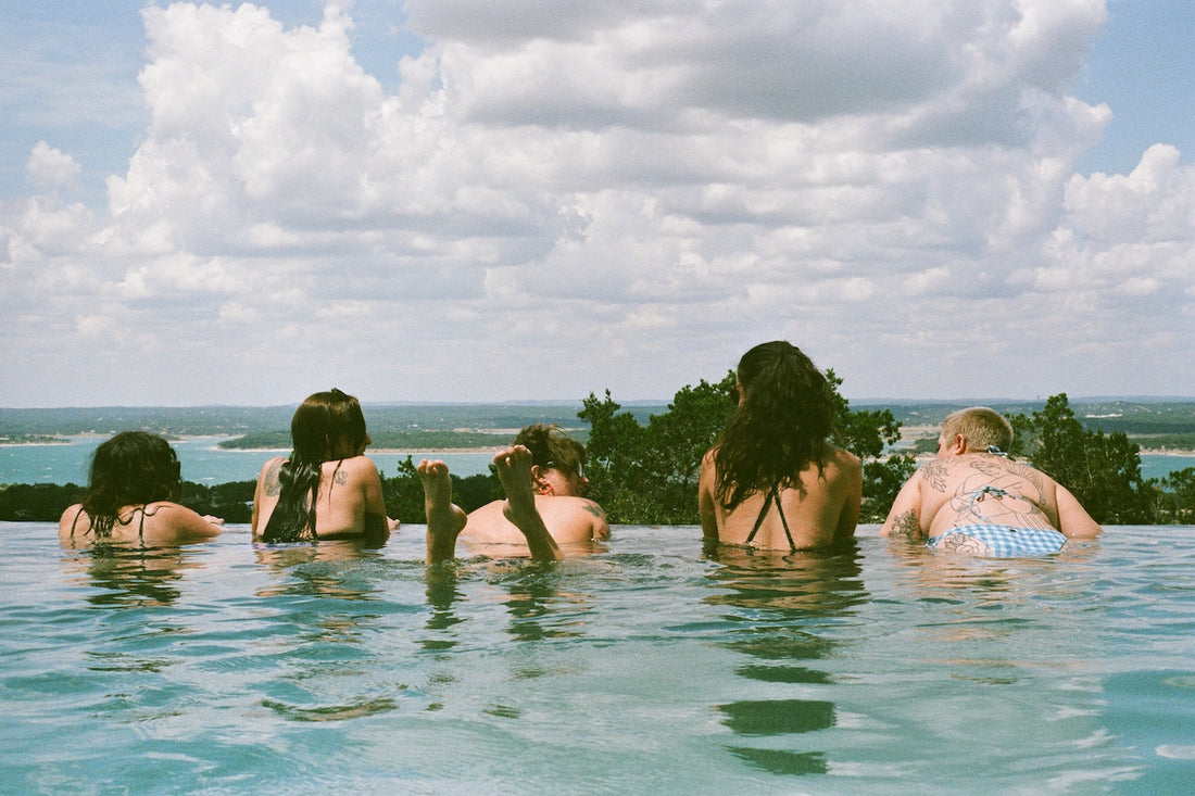 Five people in swimsuits in a pool looking out to the horizon. You can see trees, water, and sand in the distance