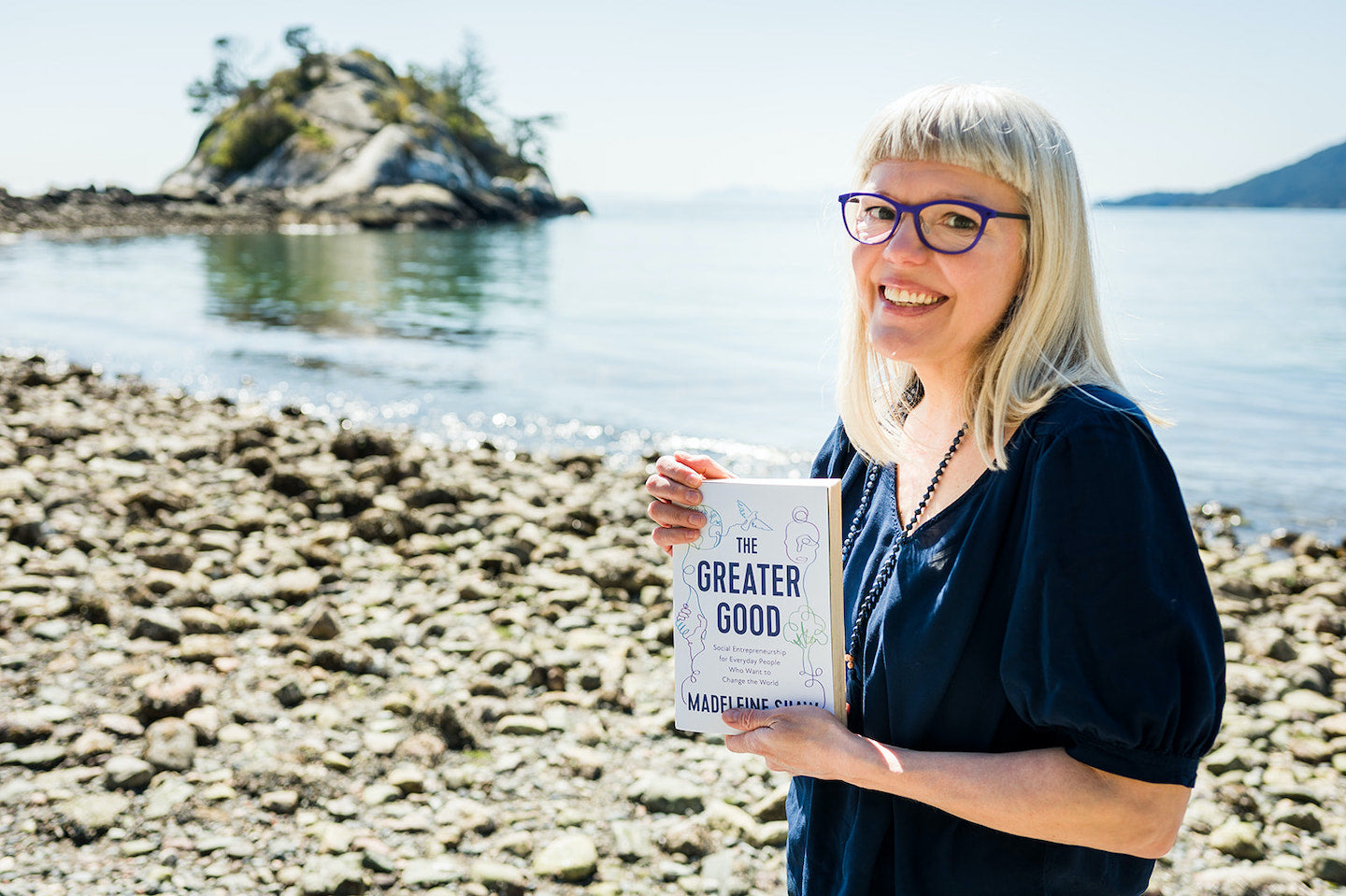 Madeleine Shaw stands on a beach holding a copy of her book, The Greater Good. She is smiling at the camera.