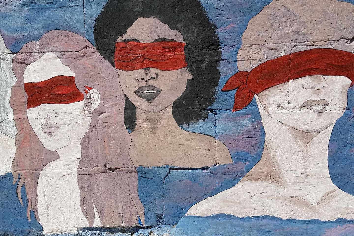 An image of a painting on a wall. The painting depicts three people, seen from the neck up, their eyes covered with a red cloth. Photo by Vulvani – www.vulvani.com