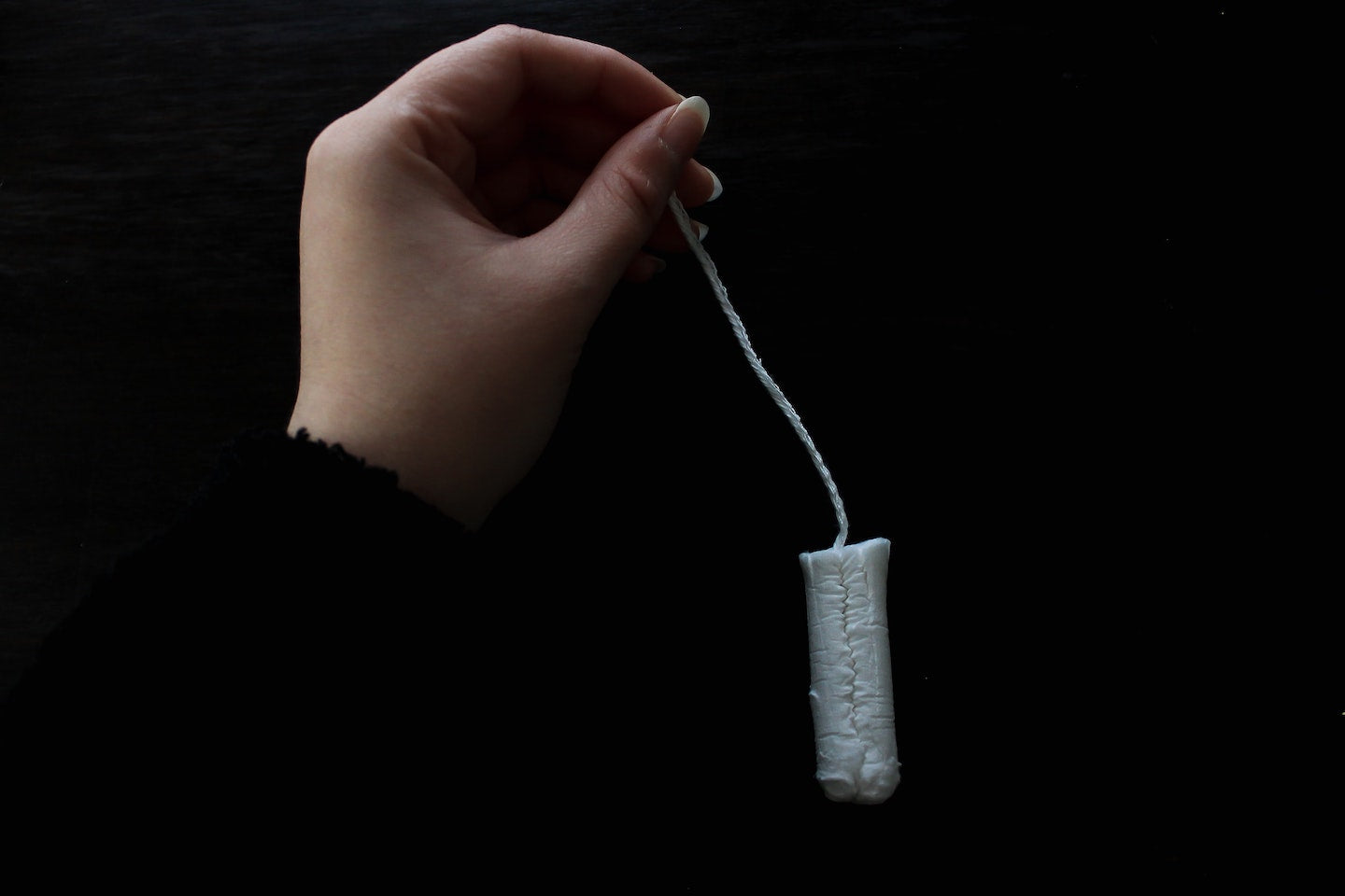 A hand holding a tampon by the string. The tampon dangles. The background is black.