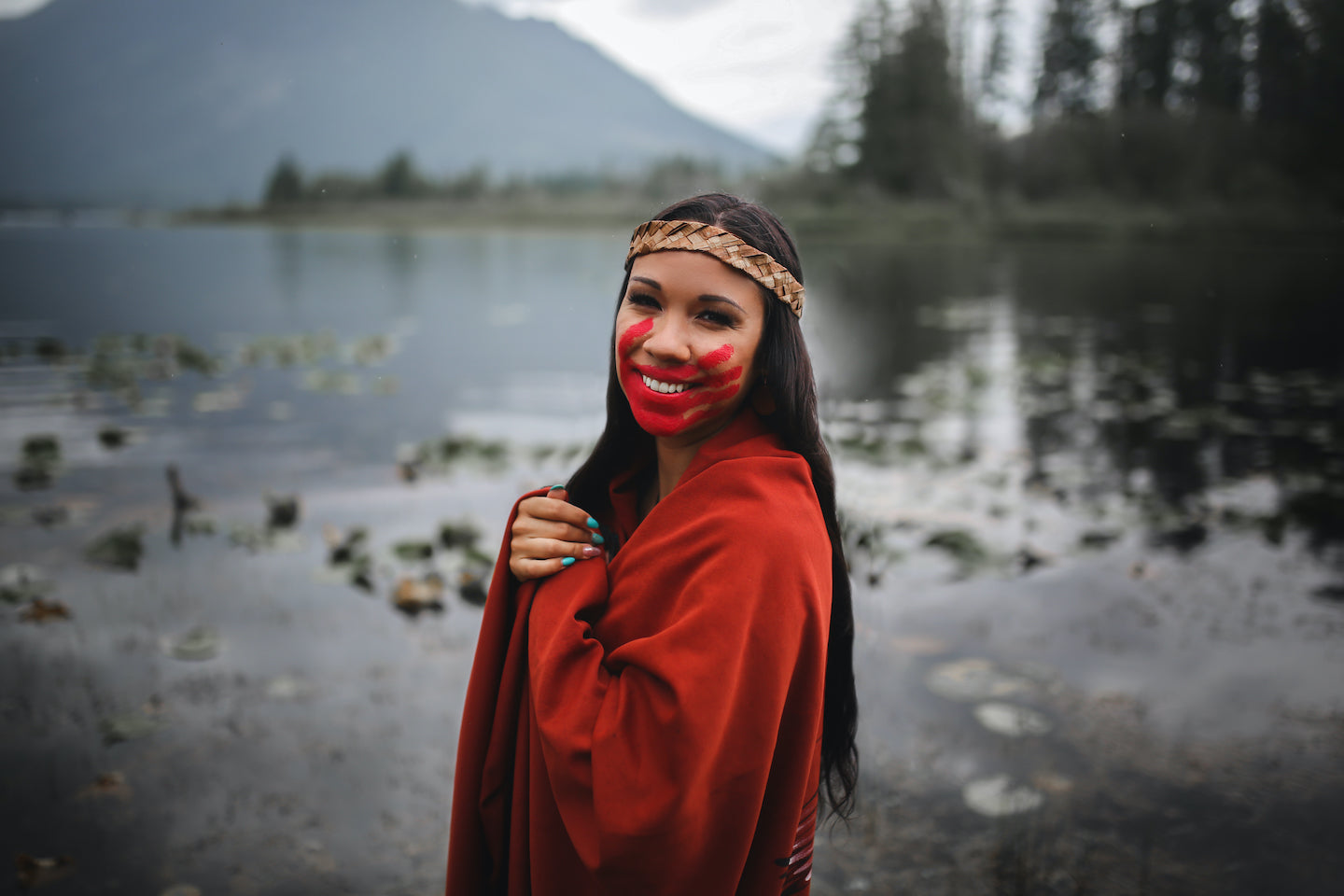Photo of Ecko Aleck standing in front of water. We can see trees and the lower part of a mountain the background. Ecko is smiling at the camera. She has a red handprint on her face, and a red robe draped over her shoulders.
