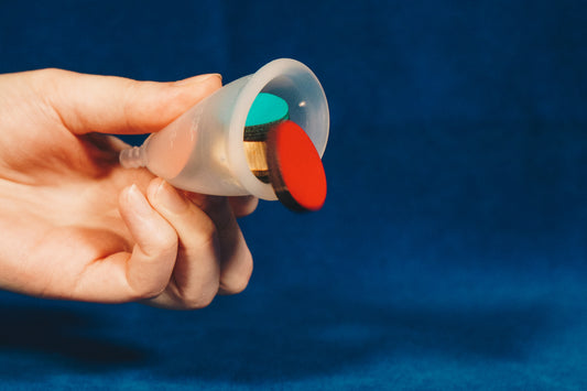 A hand holding a menstrual cup on its side. The cup is filled with coloured shapes, which have started to fall out of it. The background is blue. 