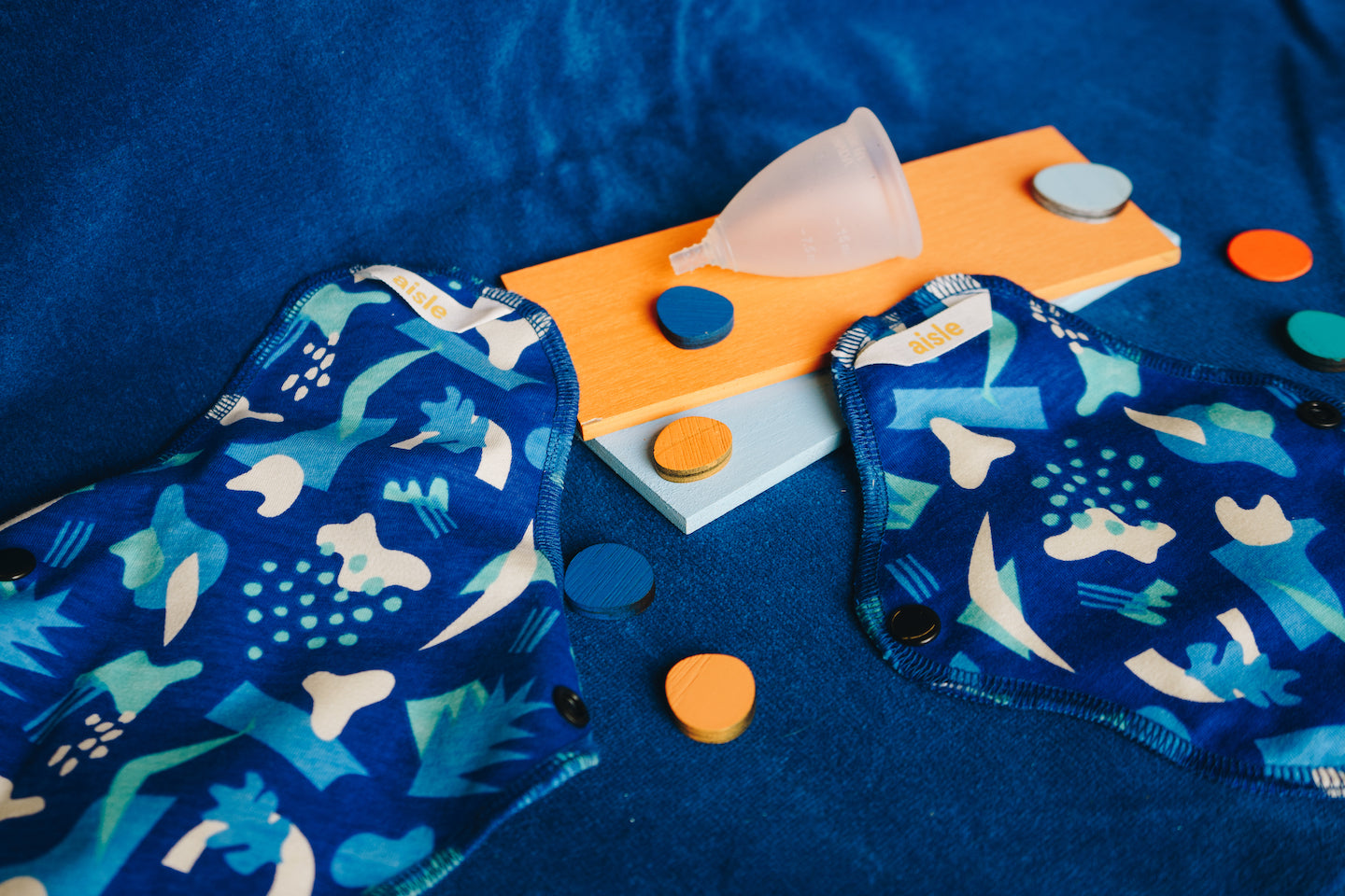 Two reusable pads and a menstrual cup placed on a blue background with various coloured shapes amongst them.
