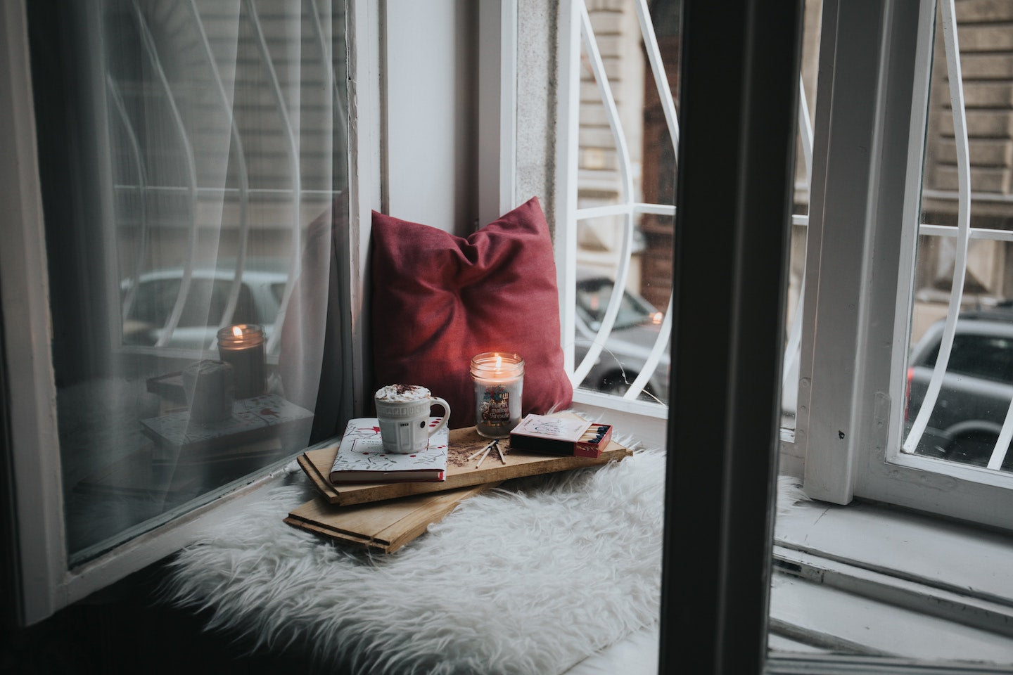 A reading nook by a window set up with a red pillow, a warm drink, a candle and some books.